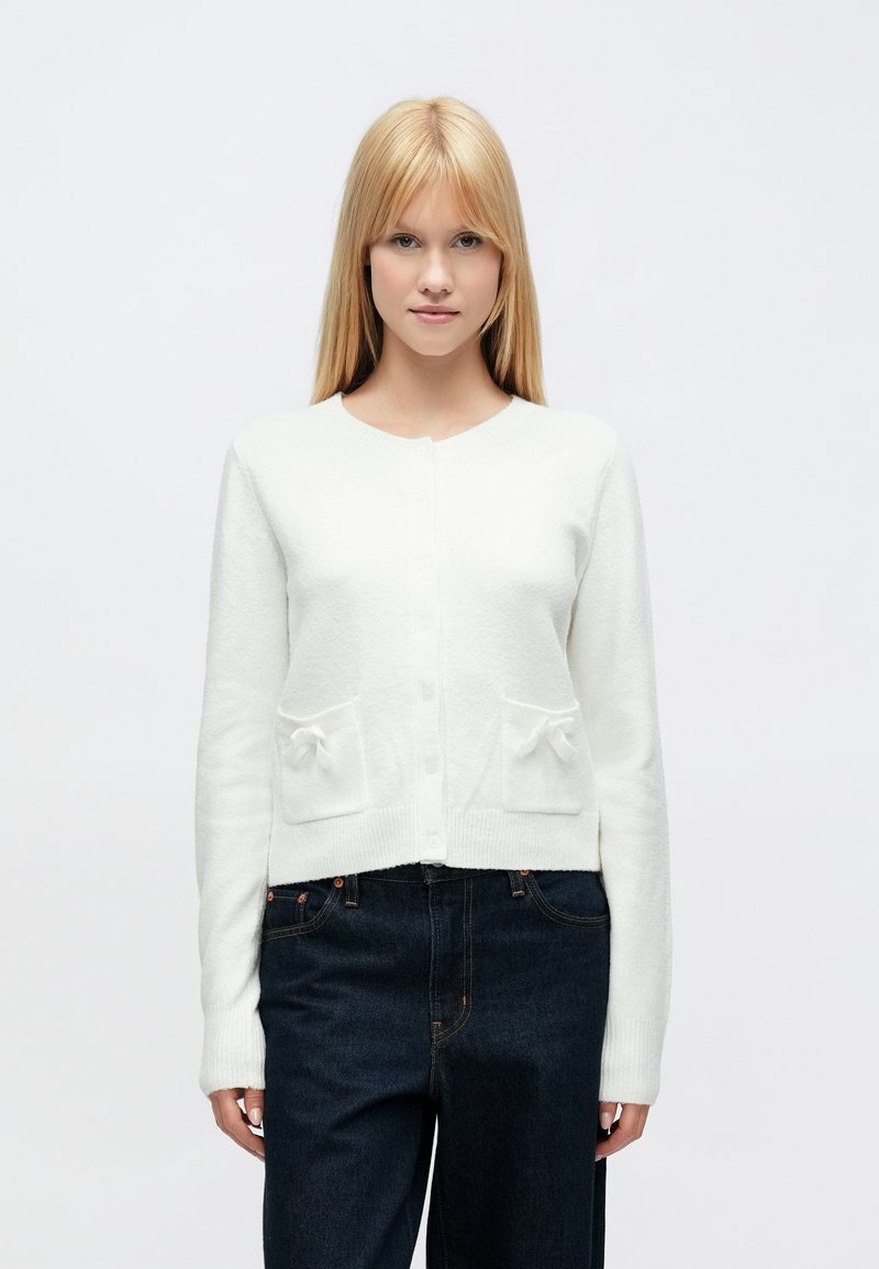 Fehér kötött kardigán hosszú ujjakkal, kerek nyakkal és két első zsebbel, amelyeken masni díszítések találhatók. Puha tapintású és rövidített fazonú.