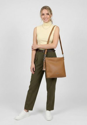 Hymyilevä nainen, joka pitää yllään hihattoman beigen topin ja oliivinväriset housut, kantaa suurta ruskeaa nahkaista olkalaukkua tupsuilla.