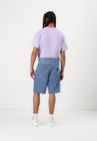 Lilac cotton T-shirt, relaxed fit, paired with blue denim shorts. Shorts feature two back pockets and a black brand patch. White sneakers visible.