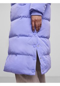 Lavendelfärgad puffjacka med horisontella quiltade sektioner, tryckknappar och ribbade ärmslut. Materialet verkar mjukt och lätt.