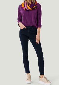 Frau trägt ein lila Oberteil mit drei Viertelärmeln, einen mehrfarbigen Schal, dunkle Skinny-Jeans und beige Sneaker und steht vor einem schlichten Hintergrund.