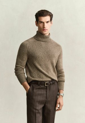 Homme portant un pull à col roulé marron et un pantalon marron à rayures fines avec une ceinture noire et une montre-bracelet argentée, debout avec une main dans la poche.