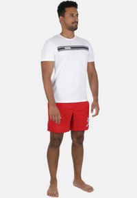 Hvid t-shirt med et sort stribet mønster og teksten "VIVID", parret med røde svømmedragter med et hvidt palmetrædesign.