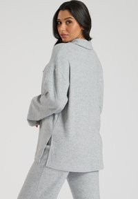 Grå sweatshirt med en afslappet pasform, lange ærmer, knaplukning ved kraven, sideslids og blød tekstur. Bæret med matchende bukser.