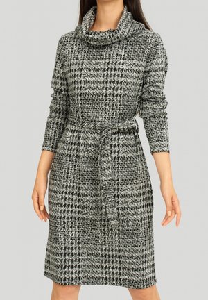 Vrouw draagt een knielange, grijs-zwarte geruite gebreide jurk met lange mouwen, een col en een bijpassende stoffen riem gestrikt in de taille.