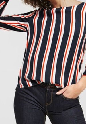 Persoon die een shirt met lange mouwen draagt met verticale marineblauwe, witte en rode strepen, gecombineerd met donkerblauwe spijkerbroek, hand rustend in de zak.