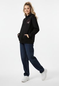 Felpa nera con cappuccio e coulisse, caratterizzata da una grafica di cuore rosso e simbolo della mano. Abbinata a jeans blu e sneaker bianche.