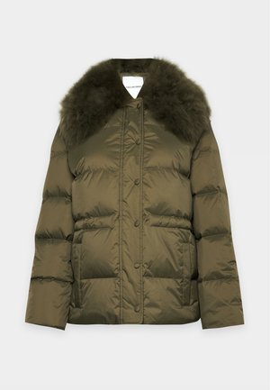 Une femme porte une veste matelassée vert olive avec une capuche bordée de fourrure foncée, un pull marron zippé, un pantalon noir et des chaussures noires épaisses.