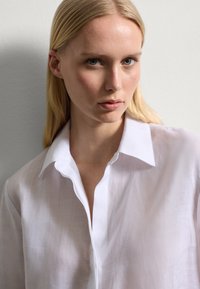 Camicia bianca, leggera, semitrasparente con colletto classico, chiusura a bottoni e vestibilità ampia; presenta una texture morbida e una leggera lucentezza.