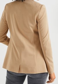 Blazer ajustado en tono beige con un acabado suave, silueta elegante y una abertura sencilla en la parte posterior. Combinado con pantalones fitted grises.