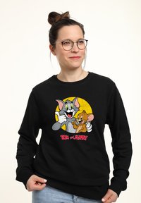 Felpa nera con un grafico dei personaggi dei cartoni animati Tom e Jerry su uno sfondo giallo, con il testo "TOM e JERRY" qui sotto.