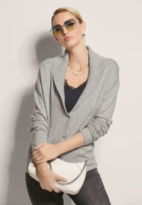 Grijze, gebreide cardigan met een sjaalkraag, knoopsluiting, gecombineerd met een zwarte top. Model houdt een witte clutch met een metalen schouderband vast.