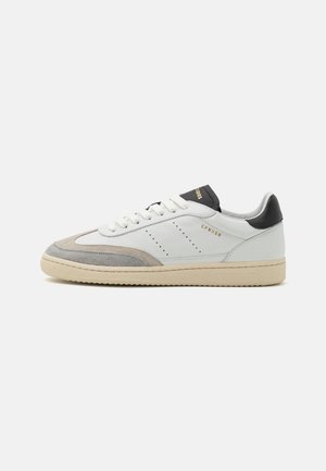 Adidași din piele albă cu accente de suede gri, vârf rotund și talpă din cauciuc texturat. Prezintă detalii perforate și toc contrastant negru.