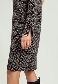 Knielange jurk in zwart en crème met een geometrisch patroon, voorzien van lange mouwen en zijspelden bij de manchetten voor meer bewegingsvrijheid.