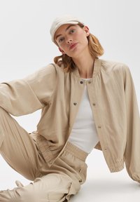 Beige bomber jacket with a ribbed collar, snap buttons, and relaxed fit, paired with matching pants and a light cream cap. Smooth texture.
