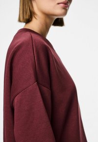 Burgunderfarget genser med senkede skuldre, rund hals og myk tekstur, sett fra siden, som viser den løse passformen og minimalistiske designet.
