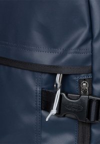 Sac à dos bleu marine en tissu texturé avec une boucle noire, des fermetures éclair et un cordon de serrage blanc. Coutures renforcées visibles.