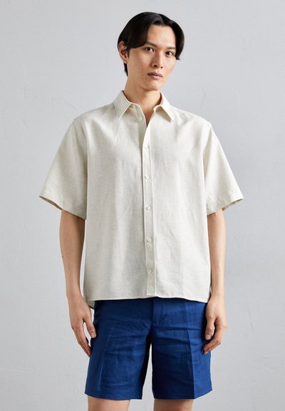 Kurzarmliges Leinenhemd in hellem Beige mit subtiler Textur, Knopfleiste und spitzem Kragen, kombiniert mit dunkelblauen Shorts.