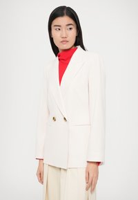 Gebroken wit dubbelgebreid colbert met gouden knopen, verlengde revers en zijzakken, gedragen over een rode coltrui. Zachte textielafwerking.