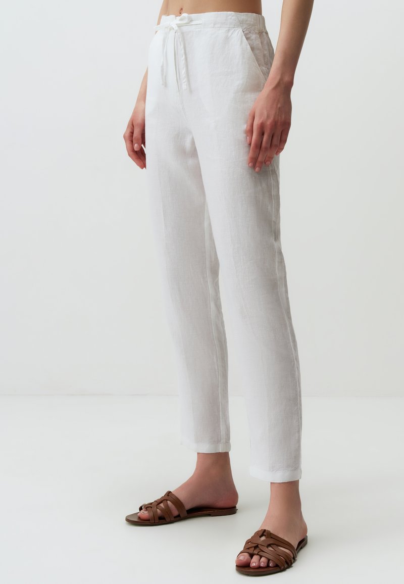 Pantalons en lin blanc avec une taille à cordon, dotés de poches et d'une coupe décontractée. Associés à des sandales brunes à lacets. Texture lisse.