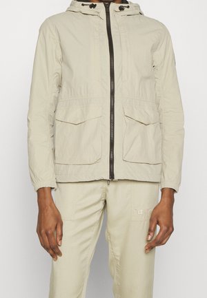 Veste à capuche beige clair avec fermeture éclair à l'avant et deux poches à rabat, portée avec un pantalon beige assorti.