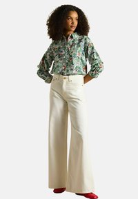 Cămașă verde cu model, cu mânecă lungă, având desene florale și geometrice, asociată cu blugi albi, cu talie înaltă și picioare largi, și încălțări roșii.