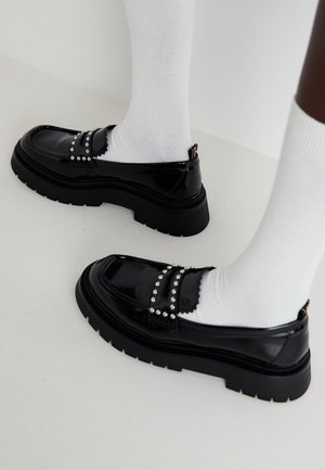 Sorte laklæder loafers med chunky såler og sølvnitter båret med hvide sokker på en lys baggrund.