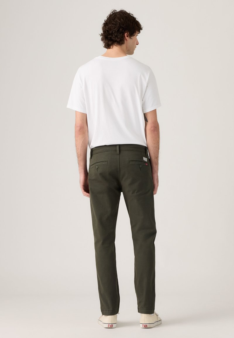 Homme aux cheveux bouclés et foncés portant un t-shirt blanc uni, un pantalon vert olive et des baskets blanches, debout de dos.