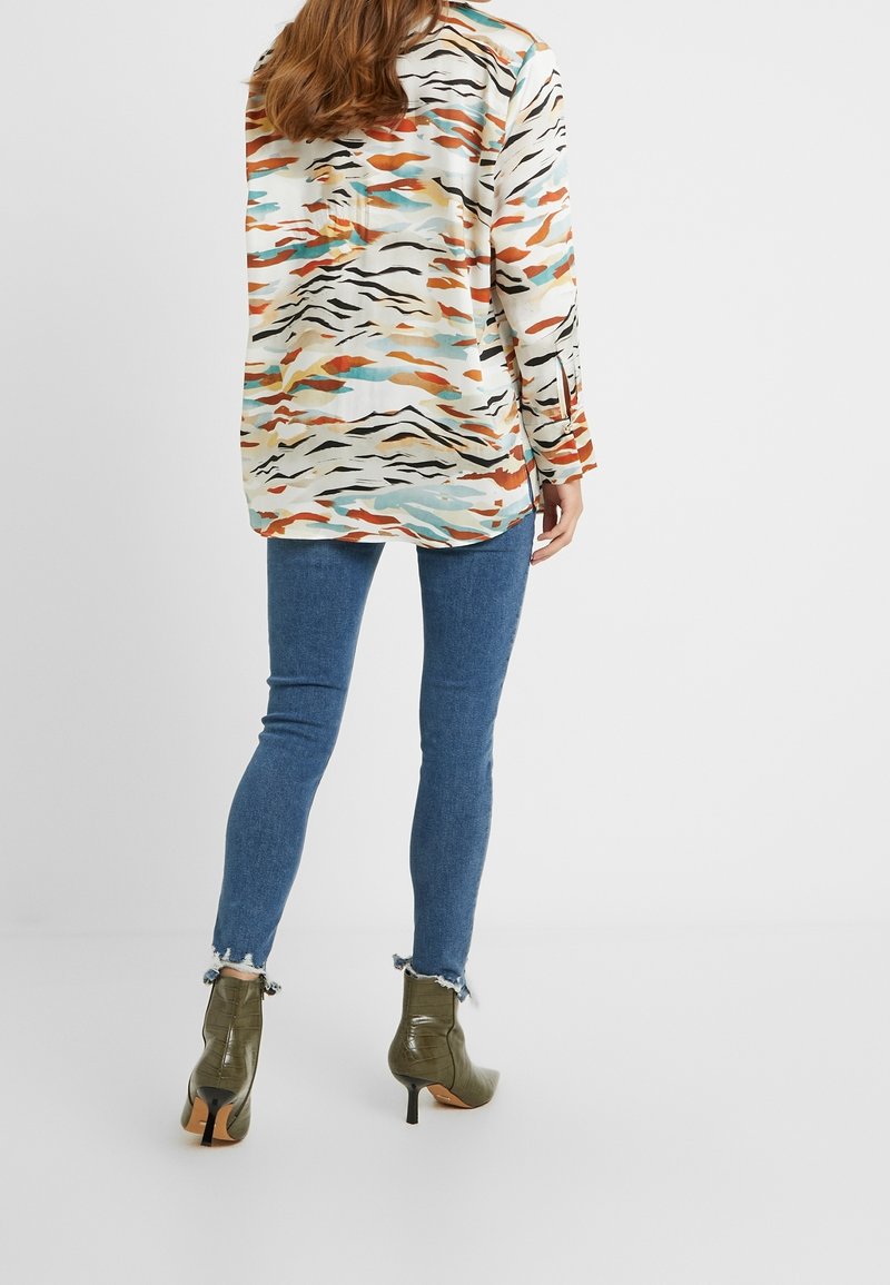 Blus med långa ärmar i ett flerfärgat abstrakt mönster med beige, orange och blå nyanser. Matchas med smala blå jeans och gröna ankelstövlar.