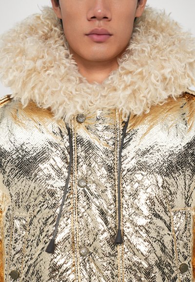 Glanzende gouden metallic parka met een gestructureerde nepbont kraag. Voorzien van zwarte trekkingkoordjes en zilveren drukkers voor sluiting.