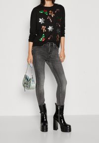 Schwarzer Pullover mit Pailletten-Gingerbread- und Bonbon-Designs, graue Jeans und glänzende schwarze Plateaustiefel. Trägt eine schillernde Handtasche.