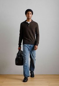 Καφέ πλέκτο πουλόβερ με γιακά, ανοιχτό μπλε τζιν παντελόνι, μαύρη τσάντα και μαύρα παπούτσια; μοντέλο περπατά σε ανοιχτό ξύλινο πάτωμα μπροστά από έναν λευκό τοίχο.