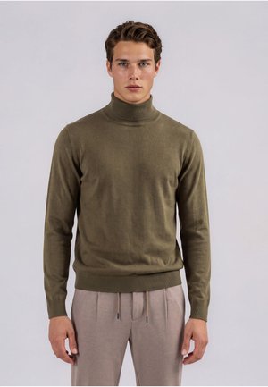 Jeune homme aux cheveux courts et bruns portant un pull à col roulé vert olive et un pantalon beige clair sur un fond uni.