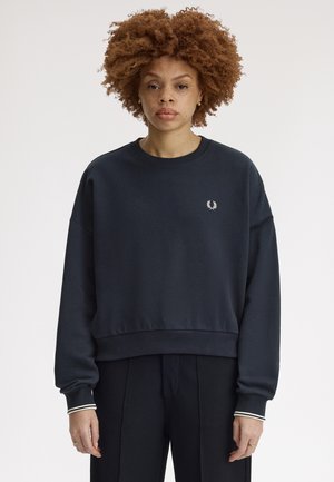 Ung kvinde med krøllet hår iført en mørkeblå sweatshirt med et lille hvidt laurbærkranslogo, stående mod en ensartet hvid baggrund.