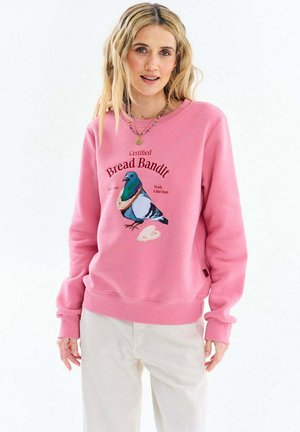 Kobieta o długich blond włosach, ubrana w różową bluzę z grafiką gołębia i chleba z napisem "Certyfikowany Złodziej Chleba" oraz jasne spodnie.