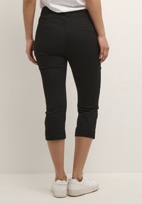 Pantalon court noir à coupe slim, avec poches arrière, ourlets retroussés et texture lisse, associé à des baskets blanches.
