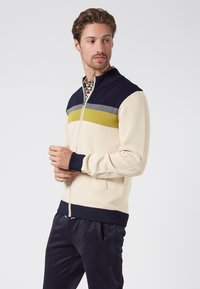 Lätt zip-up tröja i krämfärgad, med marinblå och olivfärgade ränder. Ribbad krage och mudd, slät textur med en figursydd silhuett.