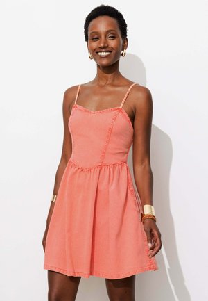 Femme souriante portant une robe corail sans manches à fines bretelles, des boucles d’oreilles créoles dorées et des bracelets dorés et orange, debout devant un fond blanc.