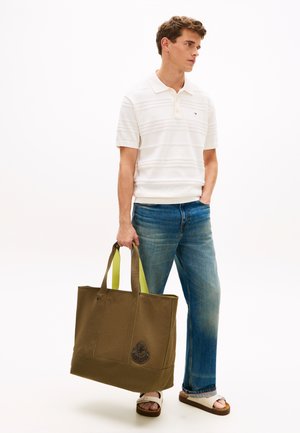 Fiatal férfi fehér pólóingben, kék farmerben és szandálban, kezében egy nagy olívazöld tote táskával, sárga fogantyúkkal és emblémával.