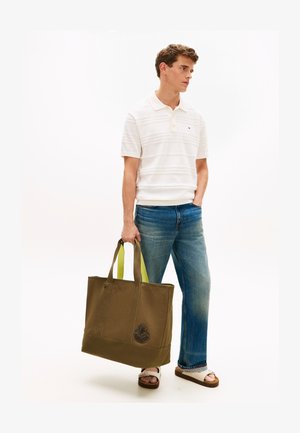 Mlajši moški v beli polo majici, modrih kavbojkah in sandalih drži veliko olivno zelene torbo za rame z rumenimi ročaji in emblemom.