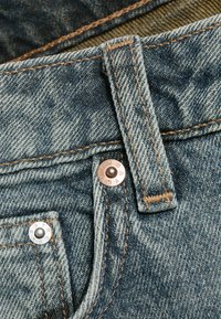 Gros plan sur un tissu en denim bleu montrant une passoire de ceinture, le bord d'une poche et deux rivets métalliques avec des lettres embossées.