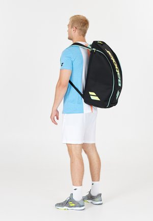 Schwarzer Tennis-Rucksack mit gelben Akzenten, geschwungenem Design, verstellbaren Trägern und Reißverschluss. Das Modell trägt ein blaues Shirt und weiße Shorts.