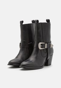 Botas de cuero negro hasta el tobillo con puntera puntiaguda, paneles elásticos acanalados y una hebilla decorativa plateada en una correa. Diseño de tacón cuadrado.