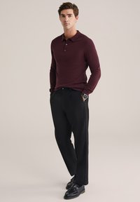 Fiatal férfi áll, kezét a zsebében tartja, bordó hosszú ujjú pólót, fekete nadrágot, fekete lakkcipőt és karórát visel.