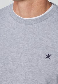 Szara bluza z okrągłym dekoltem wykonana z miękkiego materiału, z małym czarnym haftowanym logo na lewej piersi. Gładka faktura i casualowy design.