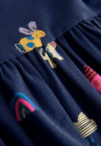 Tumši zila, fakturēta auduma kleita ar rotaļīgām izšūtām dzeltenta trusīša, kaktusa un krāsainu formu motīviem. Svārku daļa ir plandīga.