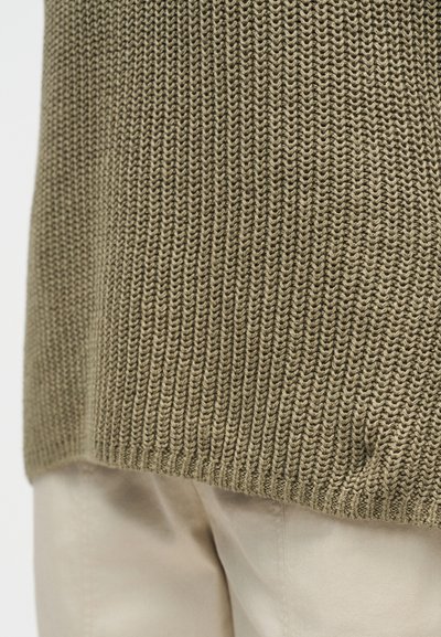 Gros plan de l'ourlet d'un pull beige en maille sur un pantalon clair, mettant en valeur le motif tricoté texturé et les détails du tissu.