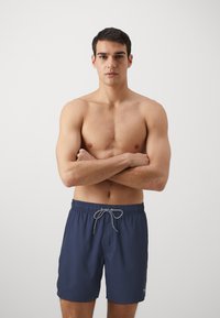 Navy swim shorts with an elastic waistband, drawstring closure, and a small logo accent. Smooth fabric texture, knee-length design.