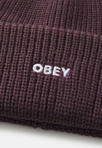 Tmavofialová pletená čiapka s ribovanou textúrou, na prednej strane s bielou vyšitou logom "OBEY". Pevný a flexibilný dizajn.