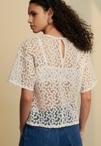 Blouse en dentelle blanche avec motif floral, manches courtes et détail d'ouverture en forme de goutte dans le dos. Texture légère, portée sur un jean bleu.